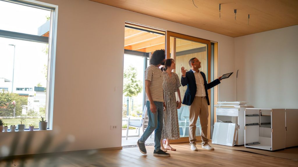A real estate agent in a blazer shows a tablet to three prospective buyers inside a bright, modern living space with large glass doors.