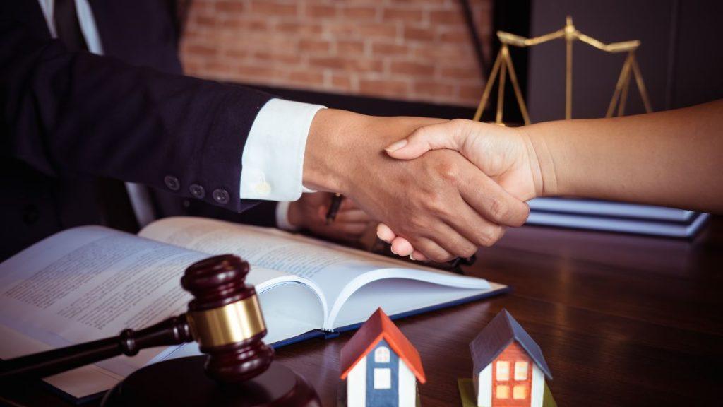 pedro-davila - Punto Critico Two people shake hands over an open contract on a desk, with a gavel and scales in the background, symbolizing a legal agreement.