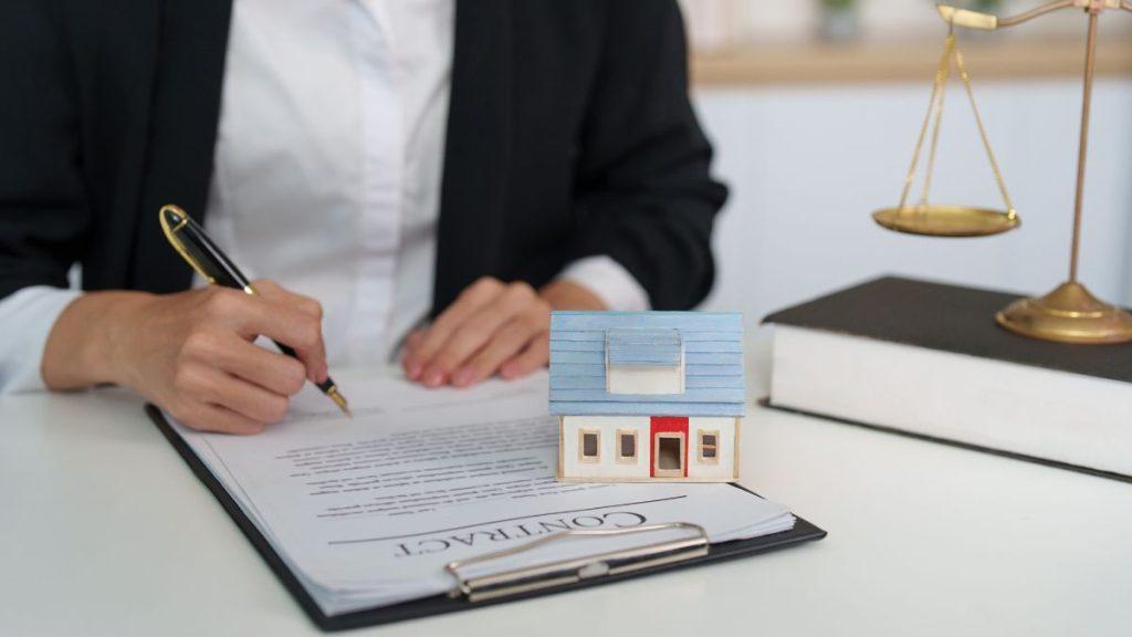 quantium-desarrollos-inmobiliaria-en-monterrey - Punto Critico Person in business attire signs a contract on a clipboard, with a small blue house model and scales of justice nearby on the desk.