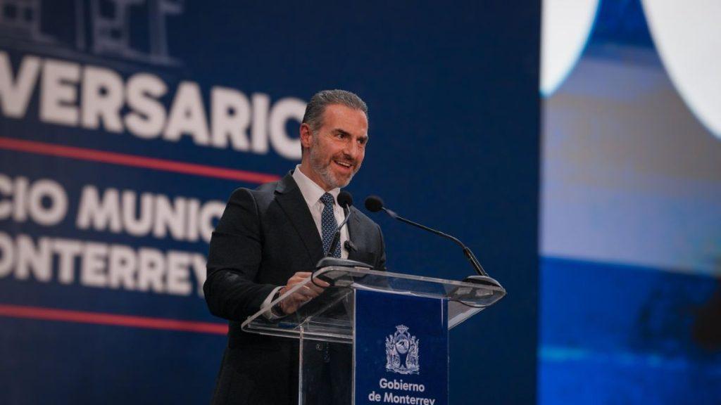Male speaker in a dark suit delivering a speech at a clear acrylic podium with a government crest, backdrop shows 'Gobierno de Monterrey' and 'ANIVERSARIO' text