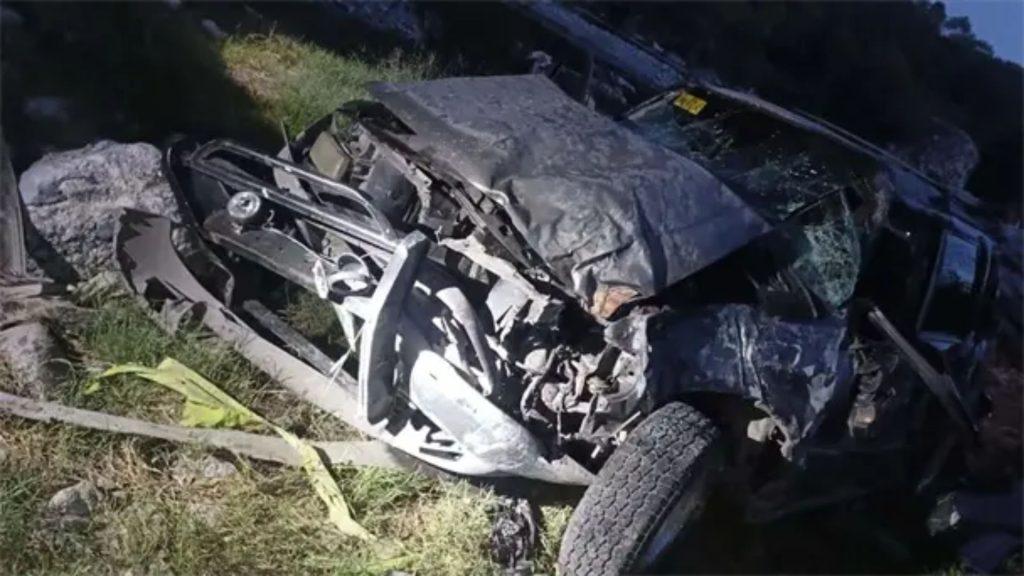 Wrecked, heavily damaged vehicle piled among rocks and grass at the roadside, exposed metal and crushed frame visible.