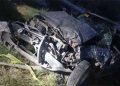 Wrecked, heavily damaged vehicle piled among rocks and grass at the roadside, exposed metal and crushed frame visible.