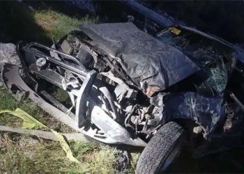 Wrecked, heavily damaged vehicle piled among rocks and grass at the roadside, exposed metal and crushed frame visible.