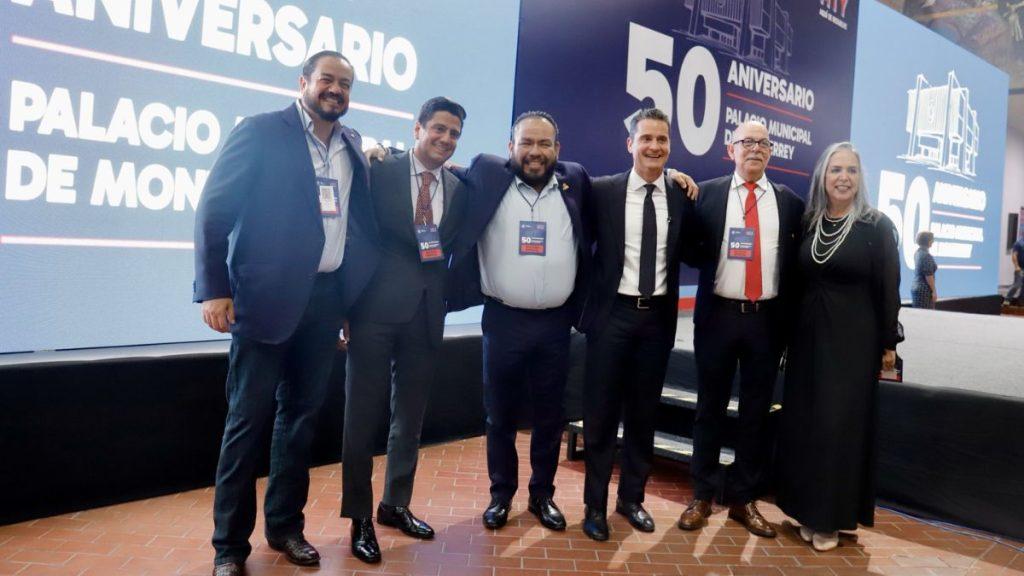 Six people in formal attire pose with arms around each other on a stage at a 50th anniversary event, badges visible on their lanyards.